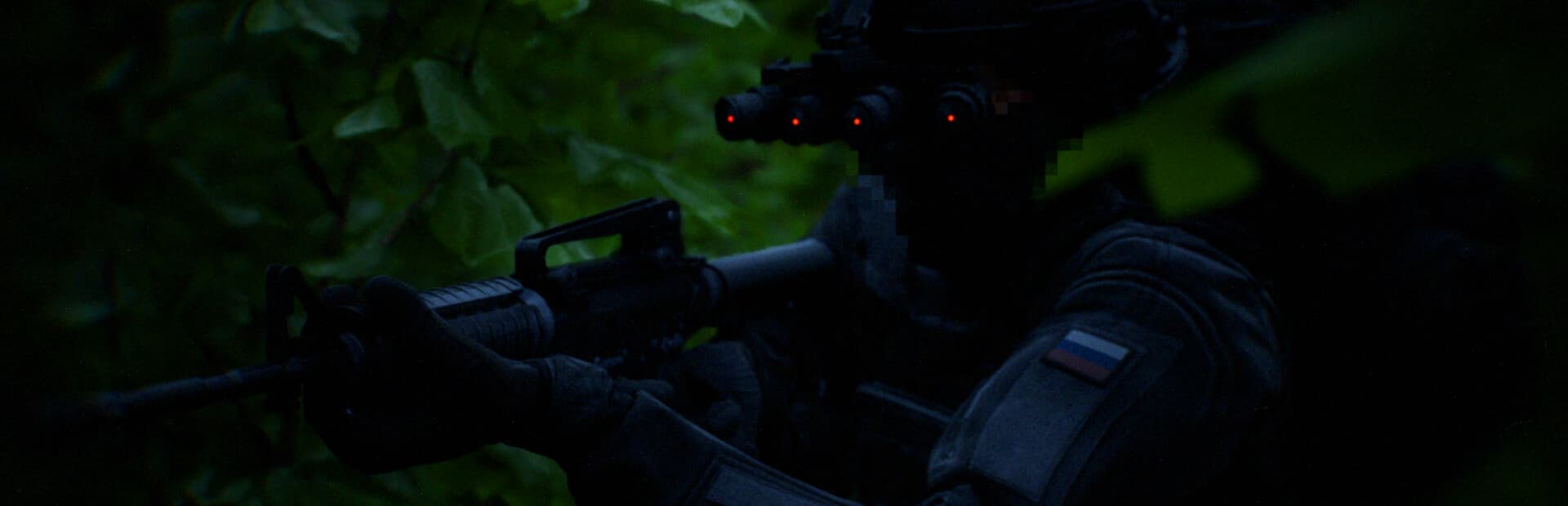 A Bodycam operator with night-vision goggles and a rifle in a dark green woodland